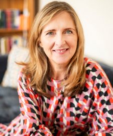 A woman (Geraldine Garrs) looking directly into the camera sitting on a sofa, wearing and red, white, pink and black checked dress.