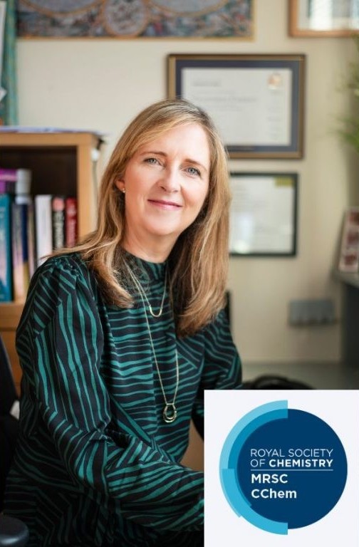 Woman in black and green striped dress sitting at a desk in front of book case and certificates